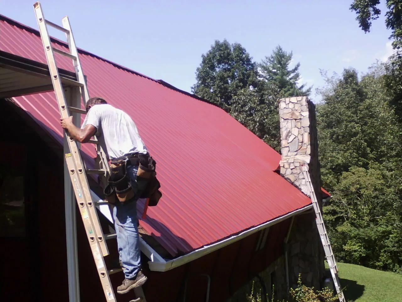 Roofer on a ladder beside a red metal roof and stone chimney — Soffit & Fascia Repair by Roofing Brothers in Bloomington Springs